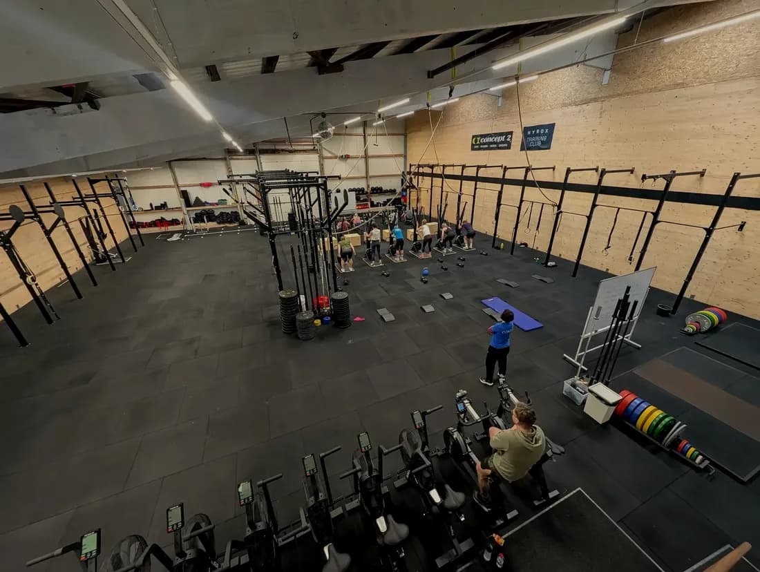 Blick in die 450 m² große Trainingsfläche von AMITY Athletics in Dülmen — Rig an der Holzwand, Concept2® Ergometer im Vordergrund, im Hintergrund eine laufende Class
